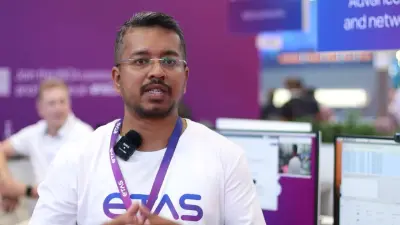 ETASのTシャツを着てマイクを手にした人物が、背景にコンピュータのディスプレイが並ぶエキシビジョンブースで話している様子。