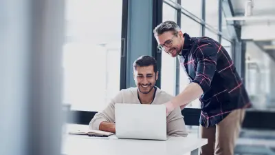 Two men collaborate on a laptop in a modern office, possibly discussing a project or sharing ideas.