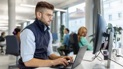 一位专注的年轻职场人士在现代开放式办公室里使用笔记本电脑工作,背景中同事们正协同合作,展现出科技驱动工作空间中的高效与创新。