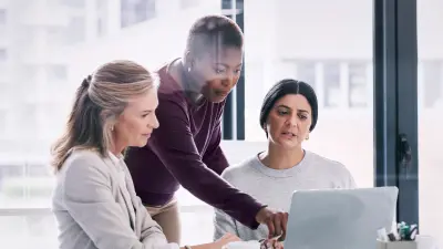 Drei Frauen schauen gemeinsam auf einen Laptop. Eine Frau zeigt mit dem Finger auf den Laptop.