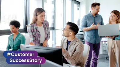 Teammitglieder arbeiten gemeinsam an Computerarbeitsplätzen in einem Büro mit dem Branding #CustomerSuccessStory.